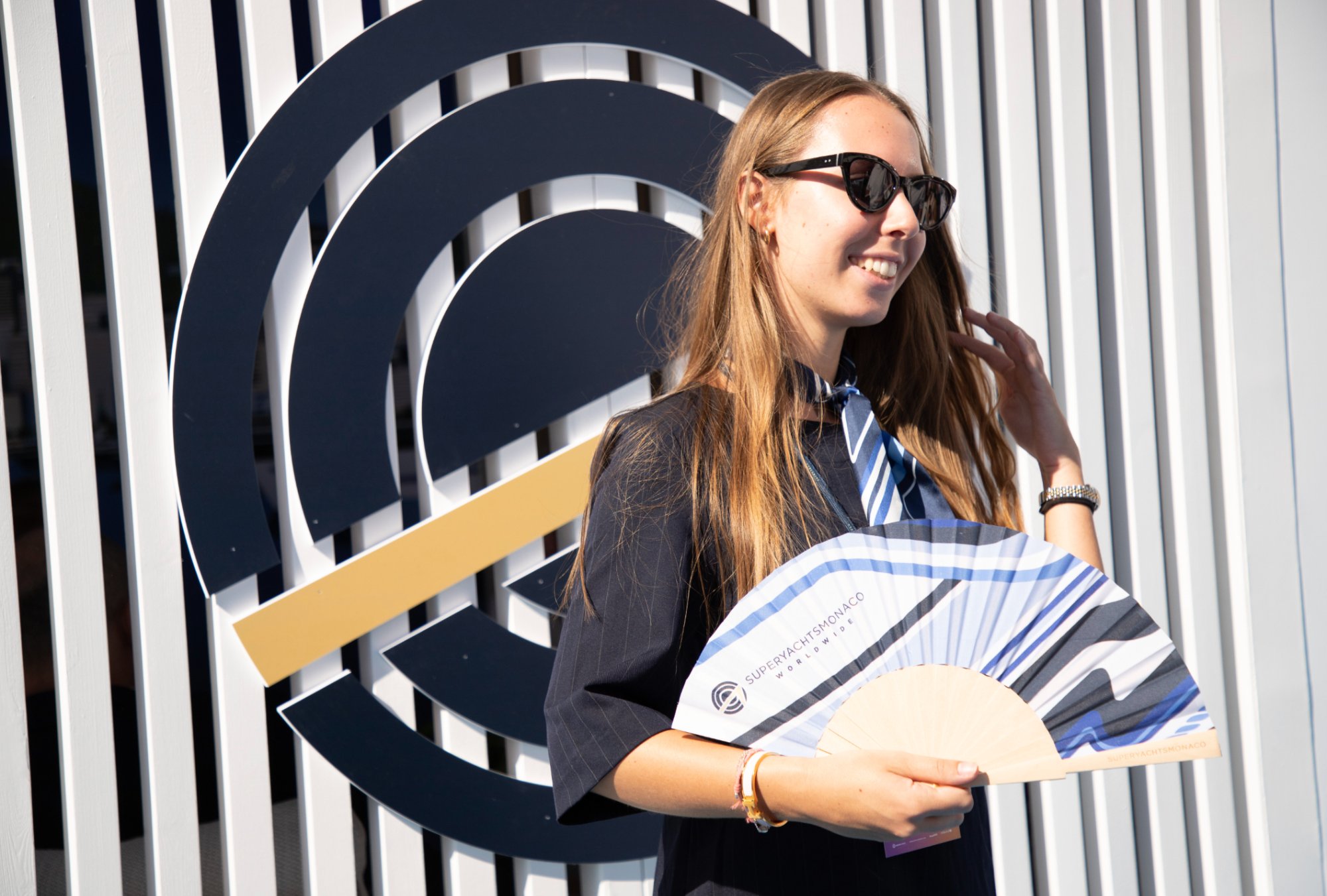 SuperYachtsMonaco team member holding branded paper fan at Monaco Yacht Show stand